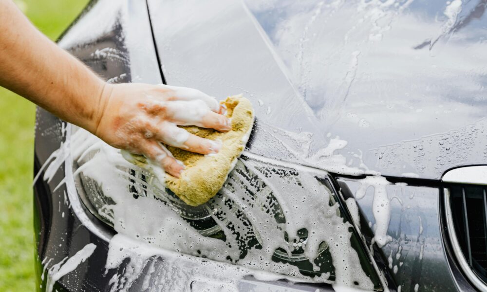 Person washing a black car's headlight with a sponge and soap outdoors.