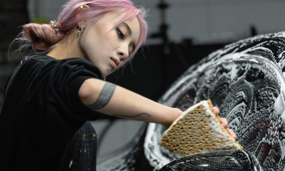 A young woman with dyed hair and tattoos cleaning a car with soap and sponge in a garage.